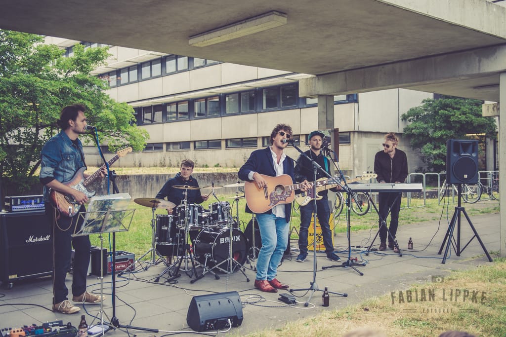 Eine fünfköpfige Band bei einem Auftritt vor einem Gebäude aus Beton