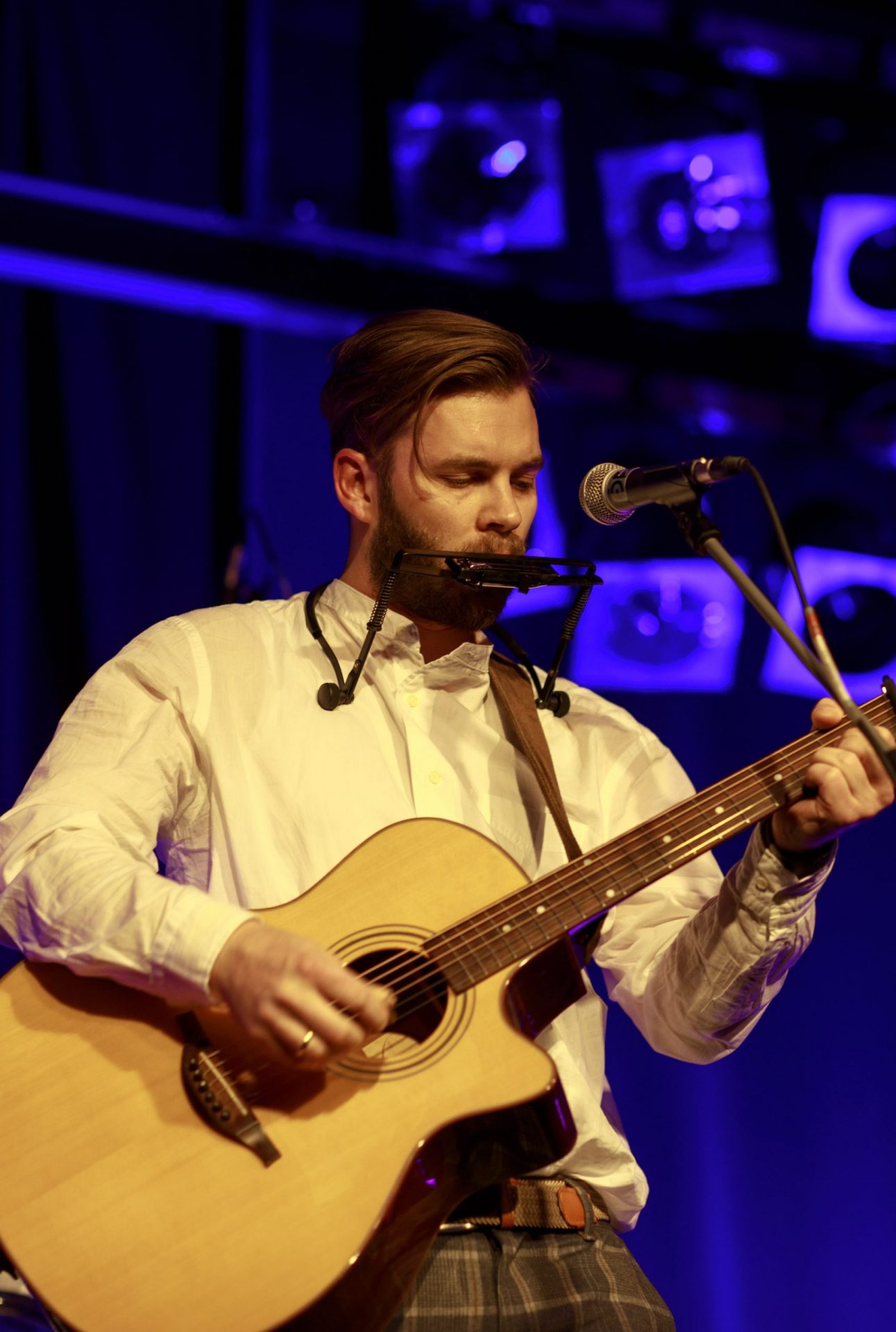 Eine Person spielt gleichzeitig Gitarre und Mundharmonika (mit Halterung) und steht auf einer Bühne.