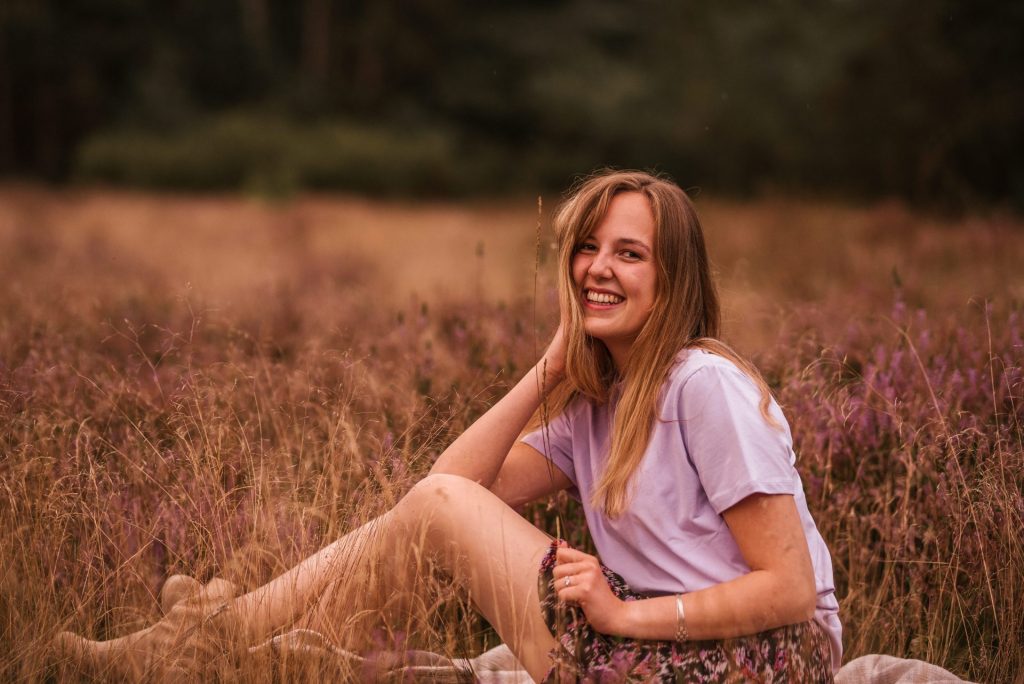 Eine Person sitzt im Gras, lächelt in die Kamera und fasst sich an den Kopf.