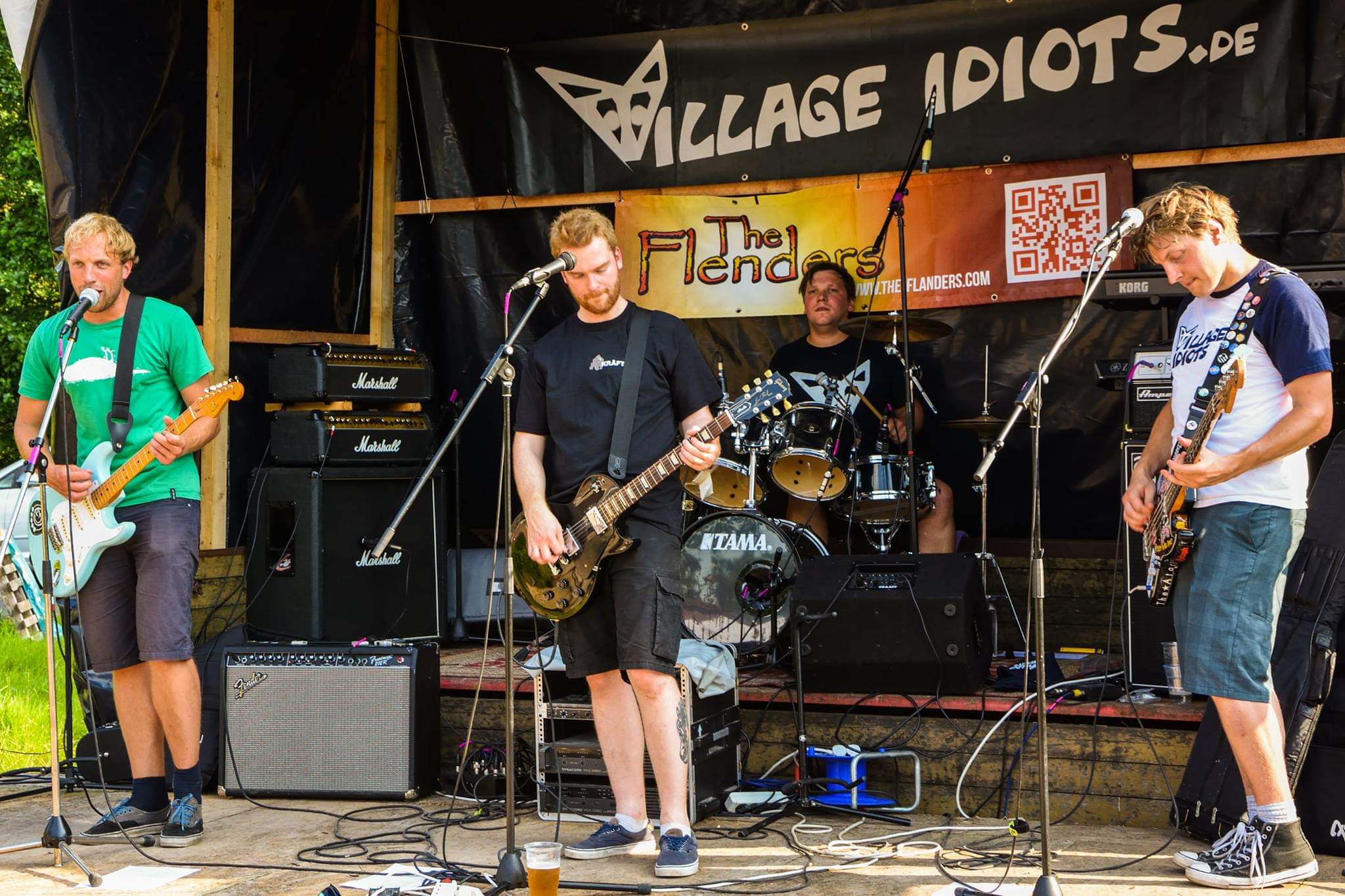 Vier Personen auf einer Bühne, zwei spielen Gitarre, eine Bass, eine sitzt am Schlagzeug. Im Hintergrund einer Banner mit der Aufschrift "Village Idiots".
