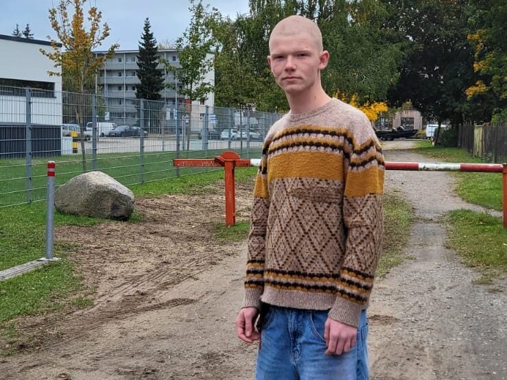 Person mit Glatze und Strickpullover vor einem Spielplatz, im Hintergrund Bäume und ein Häuserblock